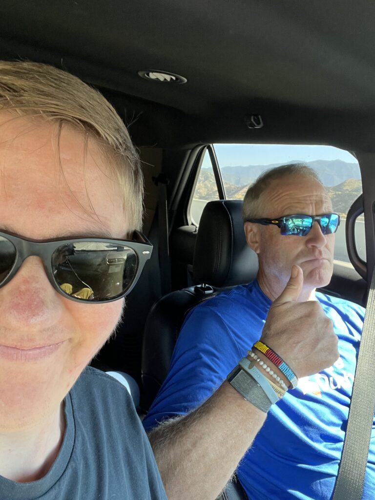 Two men pose for a selfie inside a car. The one on the right gives a thumbs up gesture.