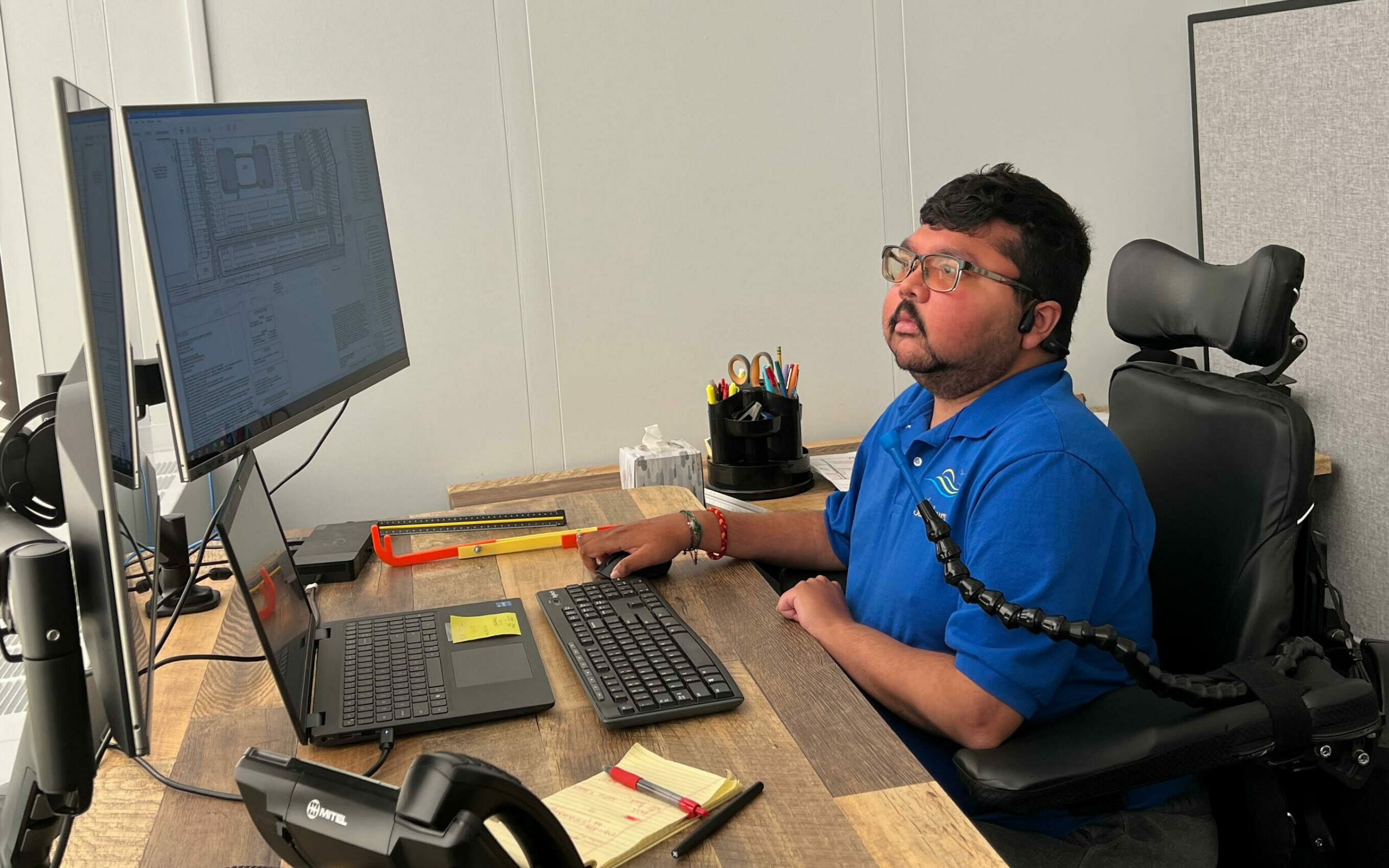 An Indian man with Duchenne muscular dystrophy in a power wheelchair sits in front of a desk with two large screens and a laptop.
