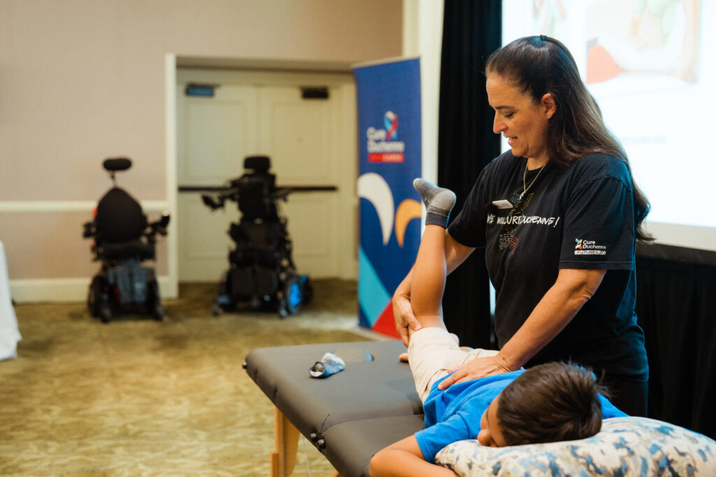 A CureDuchenne Certified physical therapist demonstrates a stretching technique on a young boy during a hands-on workshop, featuring medical equipment and educational materials in the background.